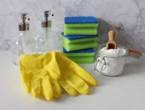 Gelbe Putzhandschuhe, zwei Glas-Sprühflaschen, Spülschwämme und ein Glas mit Natron darin.