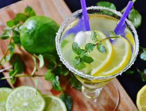 Ein Glas mit einem hellen Cocktail oder einer Limonade darin, zusammen mit Zitronenscheiben. Daneben liegen ein paar grüne Limetten.