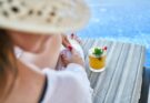 Blick von hinten auf eine Frau, die auf einem Liegestuhl am Pool sitzt. Neben ihr steht ein Cocktail.