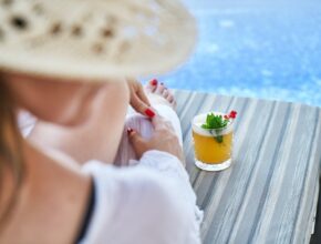 Blick von hinten auf eine Frau, die auf einem Liegestuhl am Pool sitzt. Neben ihr steht ein Cocktail.