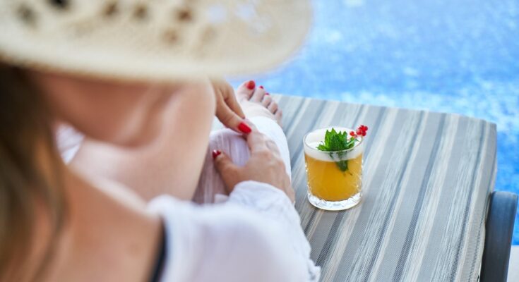 Blick von hinten auf eine Frau, die auf einem Liegestuhl am Pool sitzt. Neben ihr steht ein Cocktail.