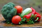 Gemüse: Ein Brokkoli, Tomaten, eine Gurke, rote Paprika und etwas Petersilie auf einem Holzbrett.