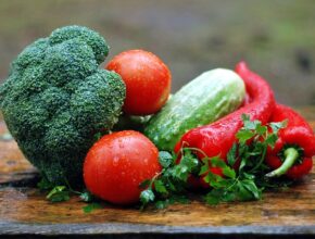 Gemüse: Ein Brokkoli, Tomaten, eine Gurke, rote Paprika und etwas Petersilie auf einem Holzbrett.