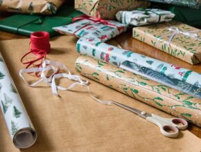 Geschenkpapierrollen auf einem Tisch. Daneben weißes und rotes Band sowie eine Schere.