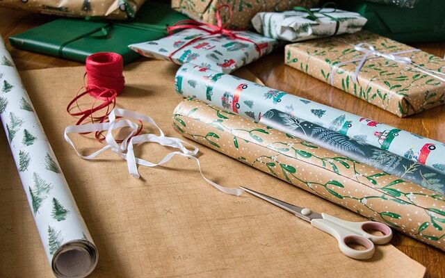 Geschenkpapierrollen auf einem Tisch. Daneben weißes und rotes Band sowie eine Schere.