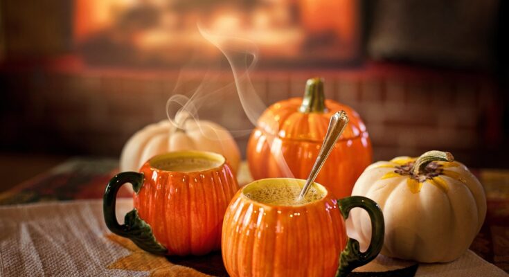 Zwei Tassen in Kürbisform mit einem Heißgetränk darin, daneben Kürbisse.