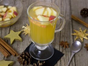 Gelber Winterpunsch in einem großen Glas. In ihm schwimmen Apfelstückchen und daneben liegen Löffel, Zimtstangen, Sternanis.