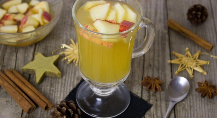 Gelber Winterpunsch in einem großen Glas. In ihm schwimmen Apfelstückchen und daneben liegen Löffel, Zimtstangen, Sternanis.