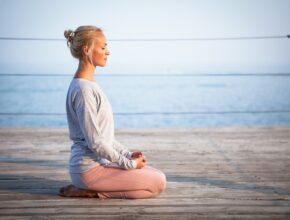 Eine meditierende Frau, die am Wasser auf einem hölzernen Untergrund sitzt.