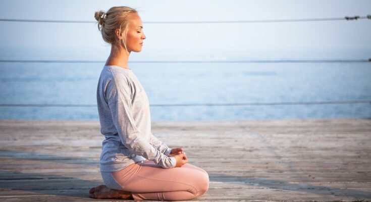 Eine meditierende Frau, die am Wasser auf einem hölzernen Untergrund sitzt.